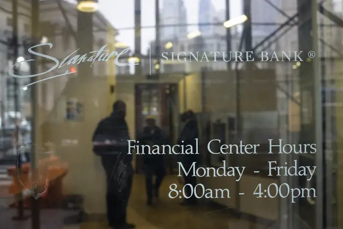 The glass doors of Signature Bank in Brooklyn shows the hours of operation as three people are huddled in the background.